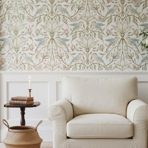 May include: A cream-colored upholstered armchair sits in front of a wall covered in wallpaper featuring a repeating pattern of light blue birds, green leaves, and pale pink flowers. A small wooden side table holds two books and a lit candle.
