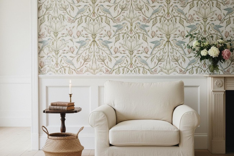 May include: A cream-colored upholstered armchair sits in front of a wall covered in wallpaper featuring a repeating pattern of light blue birds, green leaves, and pale pink flowers. A small wooden side table holds two books and a lit candle.