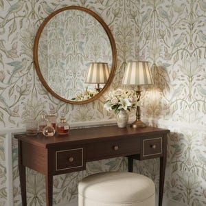 May include: A dark wood vanity table with three drawers and gold-toned knobs, paired with a round mirror in a matching wood frame. A table lamp with a striped shade and a small vase of white flowers sit on the surface, alongside three glass bottles.