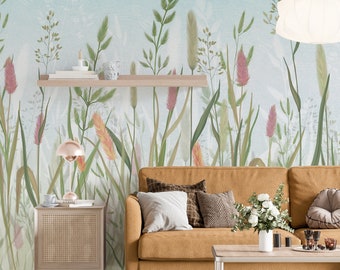 May include: A living room interior with a light blue wall featuring a floral wallpaper with tall green grasses and pink flowers. A brown couch with pillows sits in front of the wall. A wooden side table with a lamp and a coffee table with a vase of white flowers are in the room.