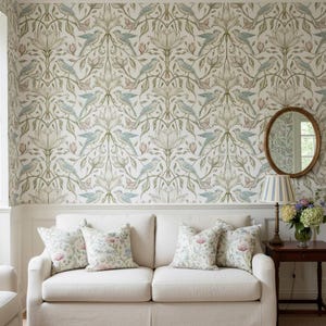 May include: A cream-colored sofa with four floral patterned throw pillows sits against a wall covered in wallpaper featuring a repeating pattern of light blue birds, green leaves, and pale pink flowers. A wooden side table holds a lamp and flowers.