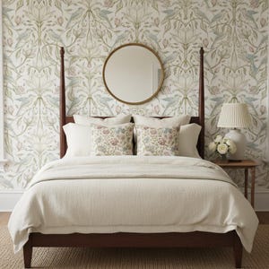 May include: A dark wood four-poster bed with a cream-colored duvet and two patterned throw pillows. Above the bed hangs a round mirror with a thin gold frame. A bedside table holds a white lamp and a small vase of white flowers.