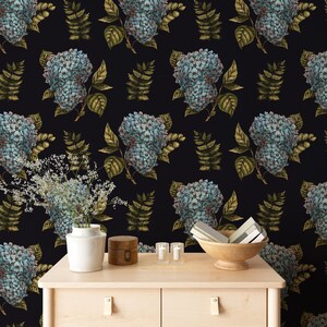 May include: A repeating pattern of blue hydrangea flowers and green fern leaves covers a dark background. A wooden cabinet holds a white vase with flowers, a small wooden box, candles, and a wooden bowl.