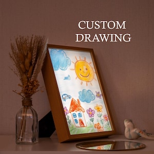 Può includere: Una cornice in legno illuminata che mostra il disegno di un bambino di una casa, un sole, nuvole, fiori e un cane. Il testo "CUSTOM DRAWING" è sopra la cornice. Un vaso con fiori secchi e un piccolo uccello decorativo sono sullo scaffale.