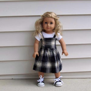Puede incluir: Muñeca con cabello rubio y ojos azules, vestida con una camiseta blanca y un vestido con peto a cuadros en azul marino, negro y crema. La muñeca también lleva zapatillas blancas con detalles negros y cordones blancos.