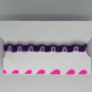 Könnte beinhalten: Ein lila und helllila Freundschaftsarmband mit einem wellenförmigen Muster auf einer weißen Karte mit rosa Herzen.