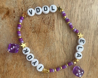 May include: Purple and gold beaded bracelet with the names "VDDL" and "ELLA" spelled out in white beads. The bracelet has two purple dice charms.