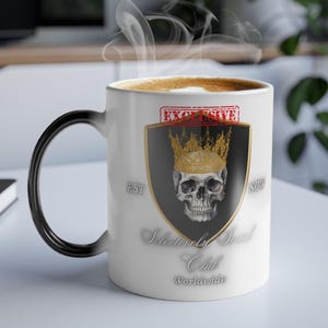 May include: White ceramic coffee mug with a black handle. The mug features a black and gold design with a skull wearing a gold crown. The text "Exclusively Skull Club Worldwide" is printed on the mug.