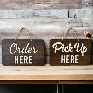 May include: Two wooden signs with white text on a brown background. The sign on the left says "Order Here" and the sign on the right says "Pick Up Here".
