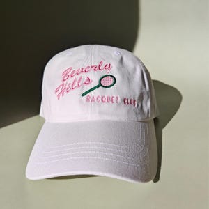 May include: White baseball cap with pink and green embroidery that reads "Beverly Hills Racquet Club" with a tennis racket graphic.