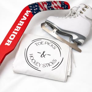 May include: A red hockey stick with the word "WARRIOR" in white, a white ice skate, and a white bag with the text "TOE PICKS & HOCKEY STICKS" in a black circle. The hockey stick has an American flag design.