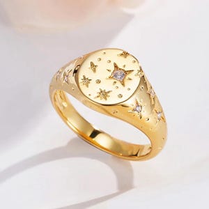 Puede incluir: Un anillo de sello dorado con un diseño celestial. El anillo presenta una cara redonda adornada con estrellas y una piedra preciosa central y transparente. Acentos en forma de estrella están engastados a lo largo de la banda, creando un accesorio elegante y brillante.