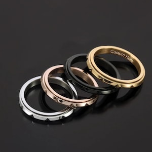 May include: Five rings in silver, rose gold, black, and gold. The gold ring has the text "Custom Here" engraved on it. Each ring has a rotating center band with a geometric design. The rings are displayed on a black surface.