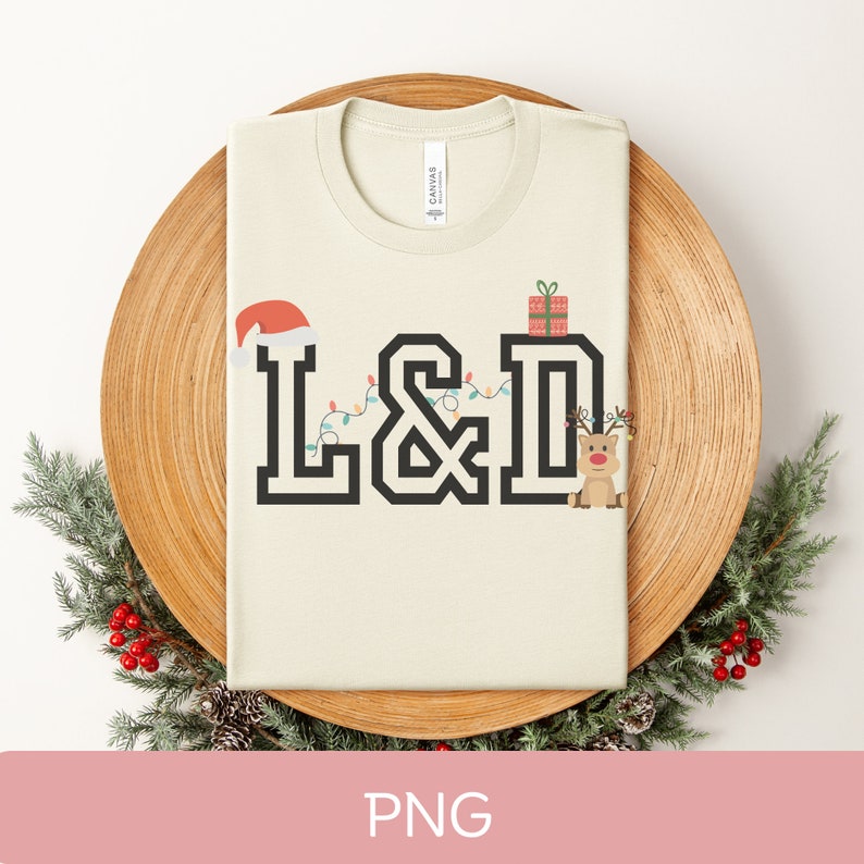 K&ouml;nnte beinhalten: Ein beiges T-Shirt mit einem schwarzen Grafikdesign der Buchstaben "L&D", verziert mit Weihnachtslichtern, einer Weihnachtsm&uuml;tze, einem Geschenk und einem Rentier. Die Buchstaben sind in einer fetten, Blockschrift.