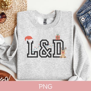 K&ouml;nnte beinhalten: Ein grauer Pullover mit einem weihnachtlichen Design, das die Buchstaben "L&D" mit einem Weihnachtsmann-Hut, Weihnachtslichtern, einer Geschenkbox und einem Rentier zeigt.