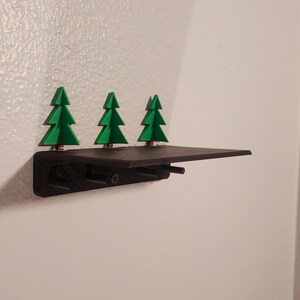 Può includere: Un ripiano a muro nero con tre ornamenti di albero di pino verde fissati sul davanti. Il ripiano è montato su una parete bianca.