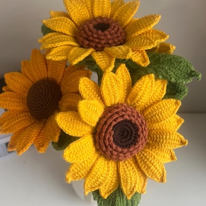 May include: A bouquet of three crocheted sunflowers. The flowers have bright yellow petals and brown centres. Green leaves are interspersed among the flowers. The sunflowers are arranged in a white vase.