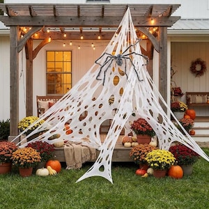 Op de afbeelding: Een groot wit decoratief spinnenweb van stof met gaten, gedrapeerd aan een houten pergola. Een grote zwarte spin is bevestigd in het midden van het web. De scène bevat oranje en gele chrysanten in potten en verschillende pompoenen.