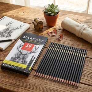 May include: A set of 14 black Markart professional sketch pencils in a rectangular box with a deer illustration, alongside a brass pencil sharpener, a sketchbook with a deer drawing, a small succulent in a terracotta pot, and a canvas roll.