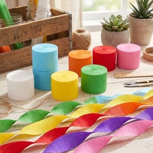 May include: A collection of crepe paper rolls in various solid colors including light blue, white, yellow, orange, red, green, and pink. Several rolls are stacked or placed upright, while long, twisted strips of the same colors extend across the surface.