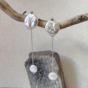 May include: A pair of drop earrings featuring iridescent white coin-shaped pearls at the top, connected by thin silver-toned metal chains to smaller, round white pearls at the bottom. The earrings have a dangling design with a metallic finish.