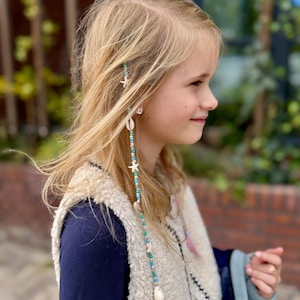 Op de afbeelding: Een jong persoon met lang blond haar, versierd met een kralen haaraccessoire. Het accessoire heeft turquoise kralen, witte schelpen en zeesterbedels. De persoon draagt een marineblauw topje en een crèmekleurig vest.