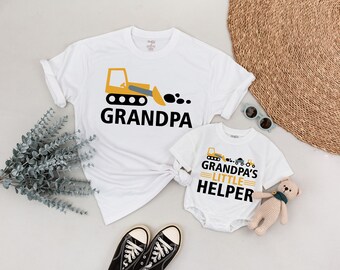 May include: White t-shirts and a onesie with construction vehicle graphics. The adult shirt says "GRANDPA" with a yellow and black bulldozer design. The onesie says "GRANDPA'S LITTLE HELPER" with a matching design.