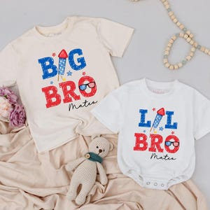 May include: Two white t-shirts with red, white, and blue firework graphics. The larger shirt says "BIG BRO" and the smaller shirt says "LIL BRO". Both shirts have the name "Mateo" printed on them.