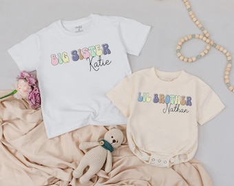 May include: Two children's shirts. One is white with "Big Sister Katie" in pastel colors. The other is a cream-colored onesie with "Lil Brother Nathan" in pastel colors. Both shirts have short sleeves.