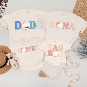 May include: A set of four beige t-shirts with a cute cartoon bao bun design. The shirts say "Doda", "Mama", "Sister", and "Bao Boo".