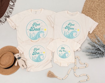 May include: Set of four cream-colored t-shirts with a blue wave and palm tree design. The shirts have the text "Rad Dad", "Rad Mom", "Rad Bro", and "The Big One" in a retro font.