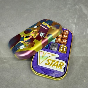 Puede incluir: Una lata del juego de cartas coleccionables Pokémon con un interior morado y dorado, que contiene dados y un soporte para cartas V STAR. La tapa de la lata presenta un diseño colorido con personajes Pokémon. Los dados están numerados.