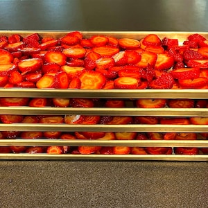 May include: Five stainless steel trays stacked on top of each other, each filled with sliced strawberries. The trays have red plastic clips on the corners.