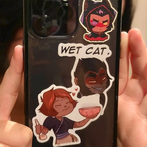 May include: A black phone case with three cartoon stickers. The top sticker is a purple-haired character with a black cat hood. The middle sticker says "WET CAT." The bottom sticker is a cartoon character with red hair holding a bowl of food.