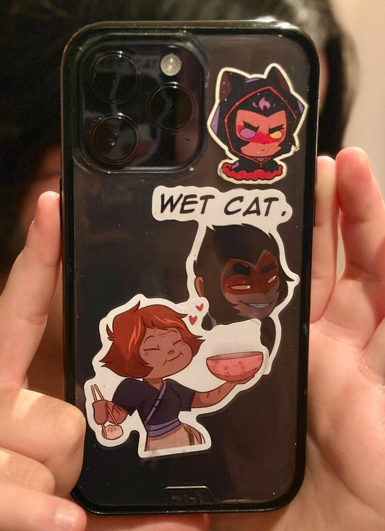 May include: A black phone case with three cartoon stickers. The top sticker is a purple-haired character with a black cat hood. The middle sticker says "WET CAT." The bottom sticker is a cartoon character with red hair holding a bowl of food.
