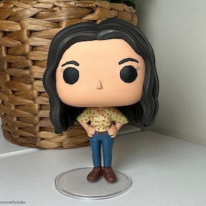 May include: A Funko Pop figure of a person with black hair, large black eyes, and a light skin tone. The figure wears a yellow shirt with red dots, blue jeans, and brown shoes. The figure is standing on a clear, circular stand.