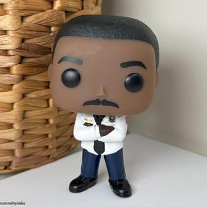 Può includere: Una statuetta in vinile Funko Pop di un uomo in uniforme bianca con cravatta nera e pantaloni blu scuro. La statuetta ha la carnagione scura, i baffi neri e i capelli neri. La statuetta è in piedi con le braccia incrociate.