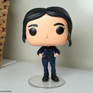 Puede incluir: Una figura de vinilo pop personalizada de una mujer con cabello negro, que lleva una chaqueta y pantalones azul oscuro. La figura tiene ojos grandes y una expresión neutral.