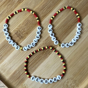Peut inclure: Trois bracelets de perles. L'un épelle "CHARLES LECLERC", un autre "LEWIS HAMILTON", et le troisième "FERRARI". Les bracelets comportent des perles rouges, noires et jaunes, et un charm en forme d'étoile dorée. Sur une surface en bois.