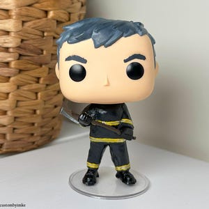 Puede incluir: Figurita personalizada de un bombero con un uniforme negro y amarillo que sostiene un hacha. La figurita tiene una cabeza grande con una cara pintada de blanco y negro y cabello negro. La figurita está de pie sobre una base de plástico transparente.