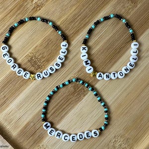 Könnte beinhalten: Drei Armbänder mit Perlen, auf denen die Namen "GEORGE RUSSELL", "KIMI ANTONELLI" und "MERCEDES" stehen. Die Armbänder haben schwarze und türkisfarbene Perlen mit goldenen Sternanhängern. Die Armbänder liegen auf einer Holzoberfläche.