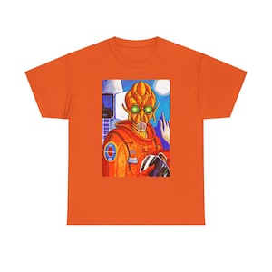 May include: Orange t-shirt with a graphic print of an alien wearing an orange spacesuit with a blue and red patch. The alien has green eyes and a toothy grin.