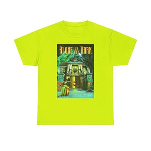 Op de afbeelding: Een neon groen T-shirt met een grafische print van een spookhuis en een persoon die een lantaarn vasthoudt. De tekst "ALONE IN THE DARK" is boven de afbeelding gedrukt.