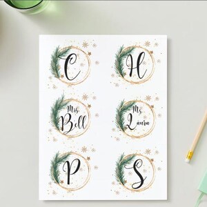 Op de afbeelding: Zes ronde, witte cadeau-etiketten met gouden glitter en groene dennentakken. De etiketten hebben de letters C, H, P, S en de namen "Mevrouw Bell" en "Mevrouw Laura" in het zwart geschreven.