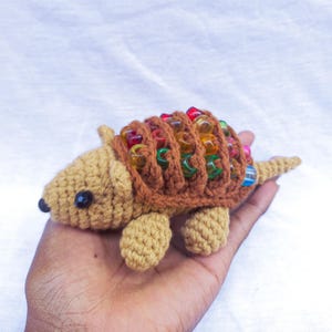 Puede incluir: Un juguete de armadillo de crochet con un cuerpo marrón y una concha hecha de cuentas de colores.