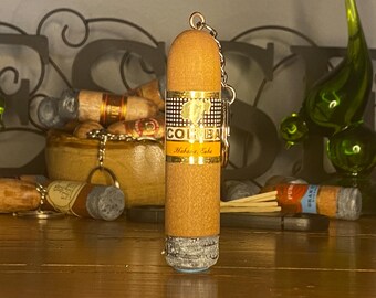 May include: A cylindrical cigar-shaped keychain featuring a tan textured body and a gold band with the black and white checkered pattern and the text COHIBA Habana, Cuba. The base of the keychain has a grey ash-like texture and a metal ring attachment.