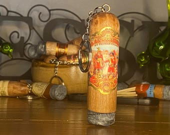 May include: A cylindrical wooden keychain shaped like a cigar, featuring a detailed label with the text 'MY FATHER CIGARS' and an illustration of historical figures. The base of the keychain has a textured gray finish resembling ash.