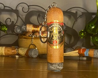 May include: A wooden keychain shaped like a cigar with a textured gray end and a gold and black band featuring a skull graphic and the text "KNUCKLE SANDWICH". The keychain includes a silver metal chain and a circular ring for attachment.