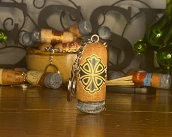 May include: A cylindrical wooden keychain featuring a black and gold cross emblem on the front. The keychain has a metallic chain and ring attached to the top, with a textured gray material at the base resembling ash, mimicking the appearance of a cigar.