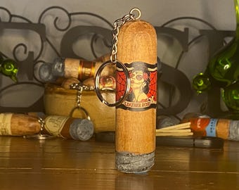 May include: A cylindrical wooden keychain shaped like a cigar with a grey textured end. The center features a label with a red spiderweb pattern and an illustration of a woman with skull face paint, with the text LEATHER ROSE printed below.
